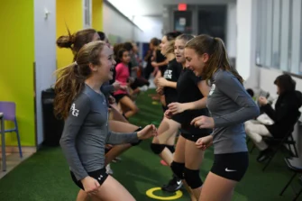 A group of young women in sportswear laughing and exercising indoors on a turf floor.