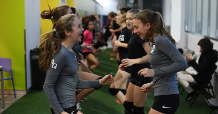 A group of young women in sportswear laughing and exercising indoors on a turf floor.