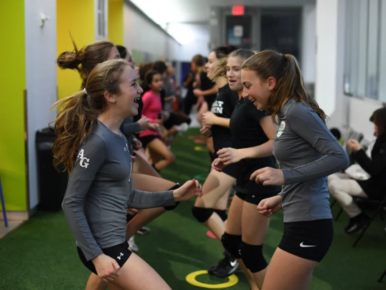 A group of young women in sportswear laughing and exercising indoors on a turf floor.
