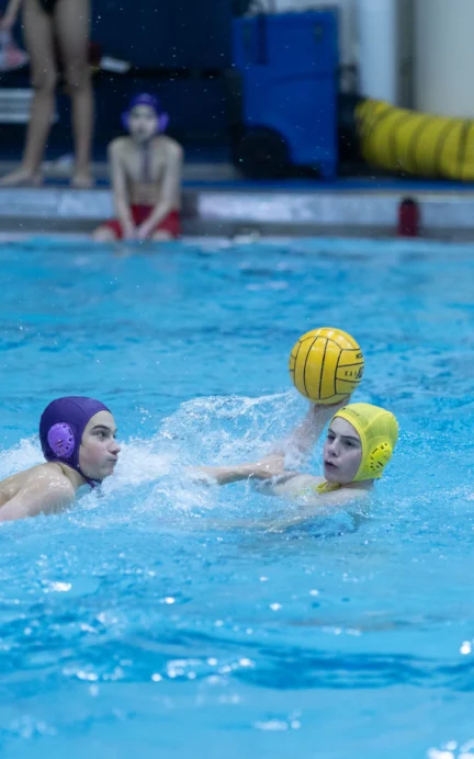 Two water polo players in action; one in a purple cap defends against another in a yellow cap holding the ball.
