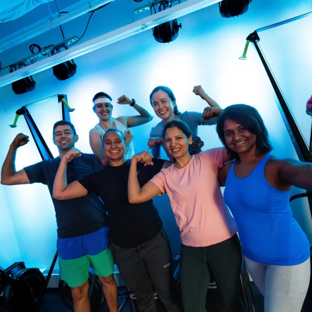 Six people in athletic clothing pose in a gym, smiling and flexing their arms after a group exercise session, with exercise equipment and blue lighting in the background.