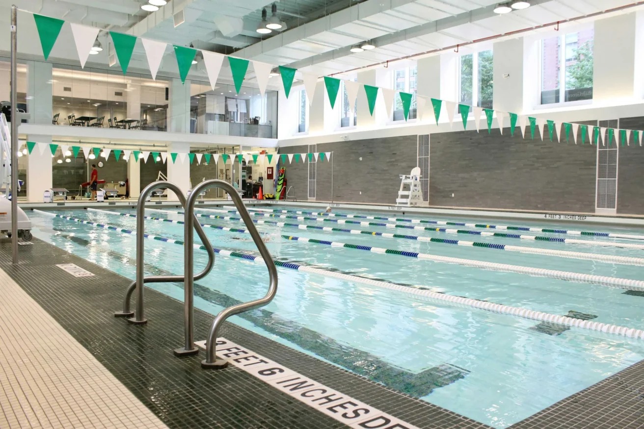 Indoor swimming pool with lane dividers and green-white pennants. Deck has a ladder and signs indicating water depth. Large windows are visible in the background.