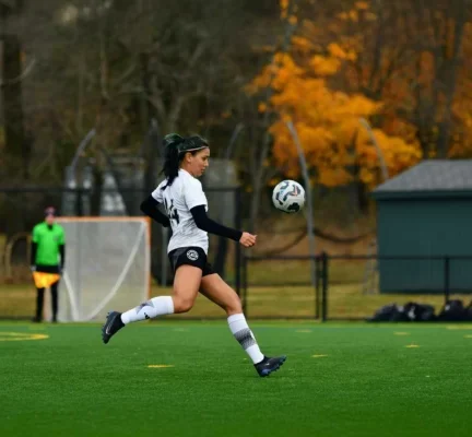 Soccer player in a white and black uniform runs with a soccer ball on a green field. Autumn trees and a goalpost are in the background.
