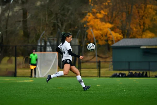 Soccer player in a white and black uniform runs with a soccer ball on a green field. Autumn trees and a goalpost are in the background.