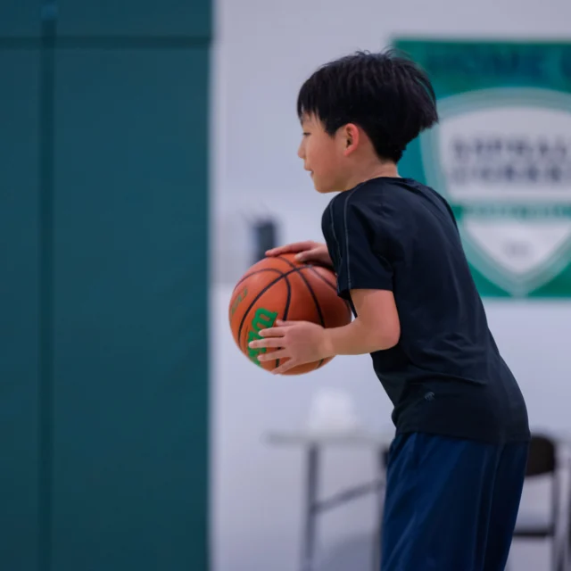 A boy holding a basketball stands indoors, ready to play. A green and white school banner is visible in the background.