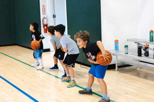 Four children lined up on a basketball court, each holding a basketball and preparing to dribble in an Asphalt Green Basketball beginner class.