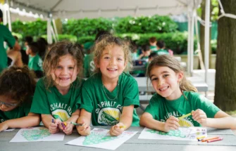 Three children at a summer camp sit at a picnic table, smiling and coloring activity sheets. They wear matching green camp t-shirts under a canopy with others in the background.