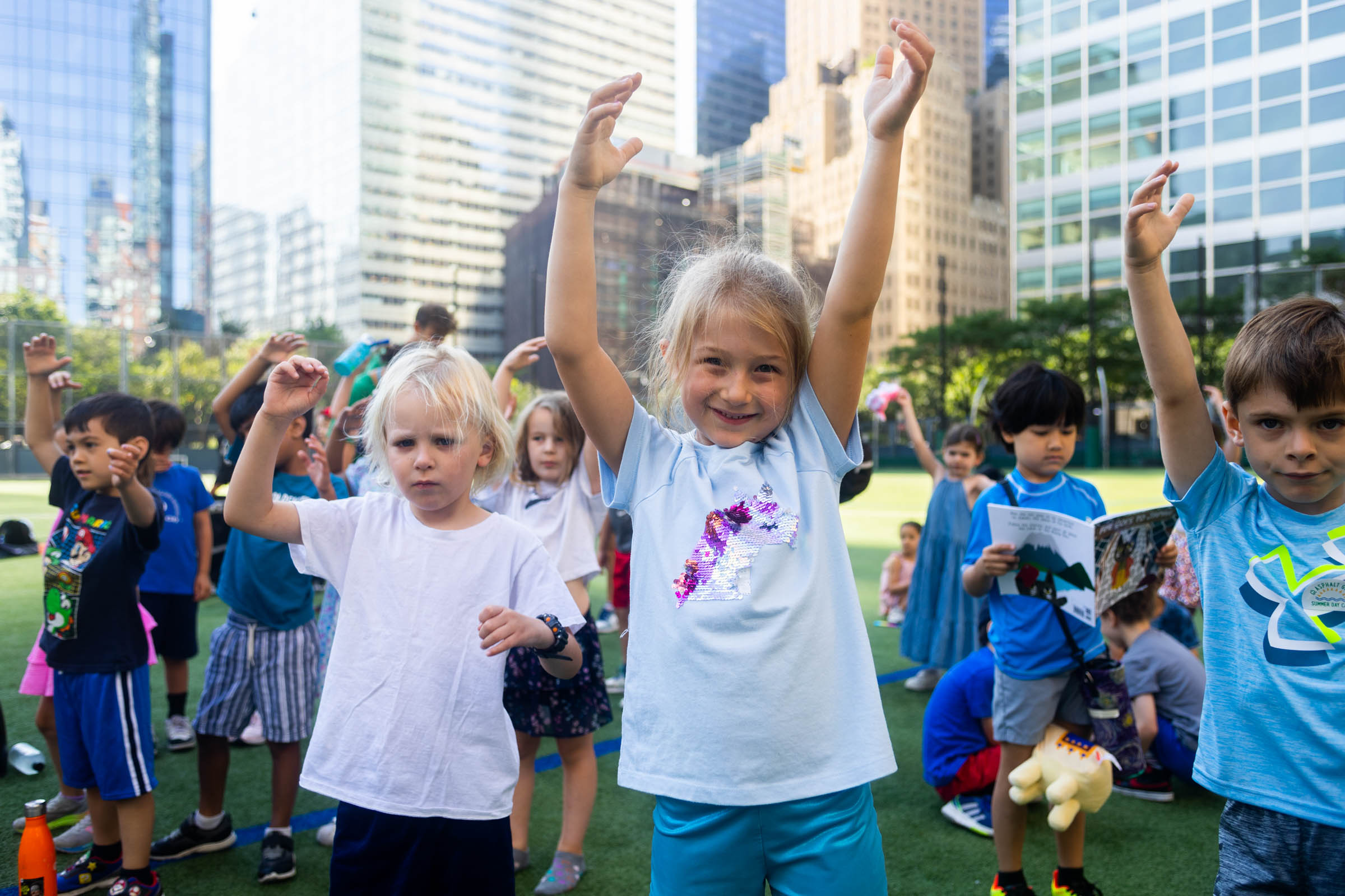 Children stand on a grassy field with their arms raised. Buildings and trees are visible in the background.