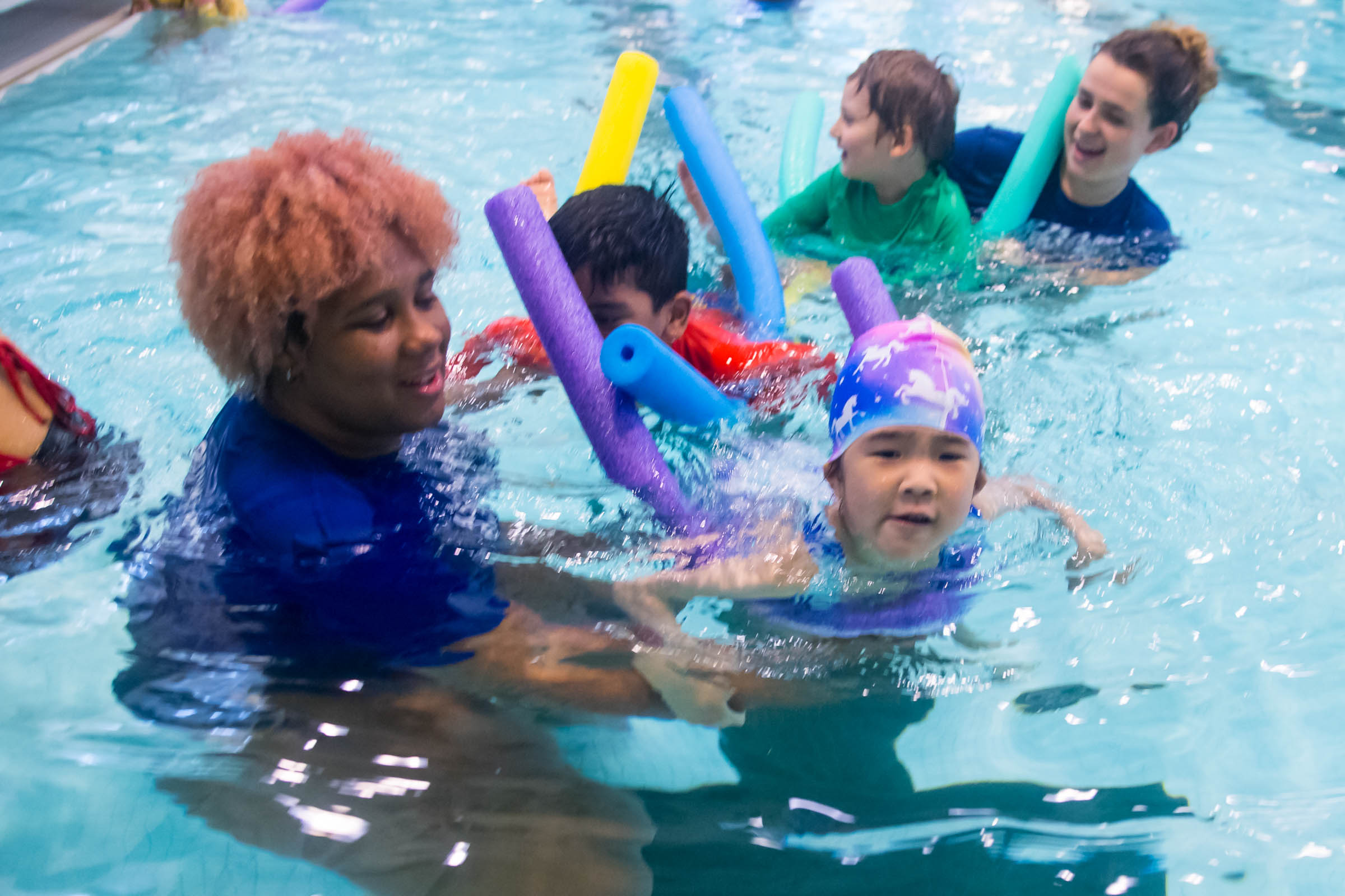 Children and adults swim in a pool with colorful pool noodles.