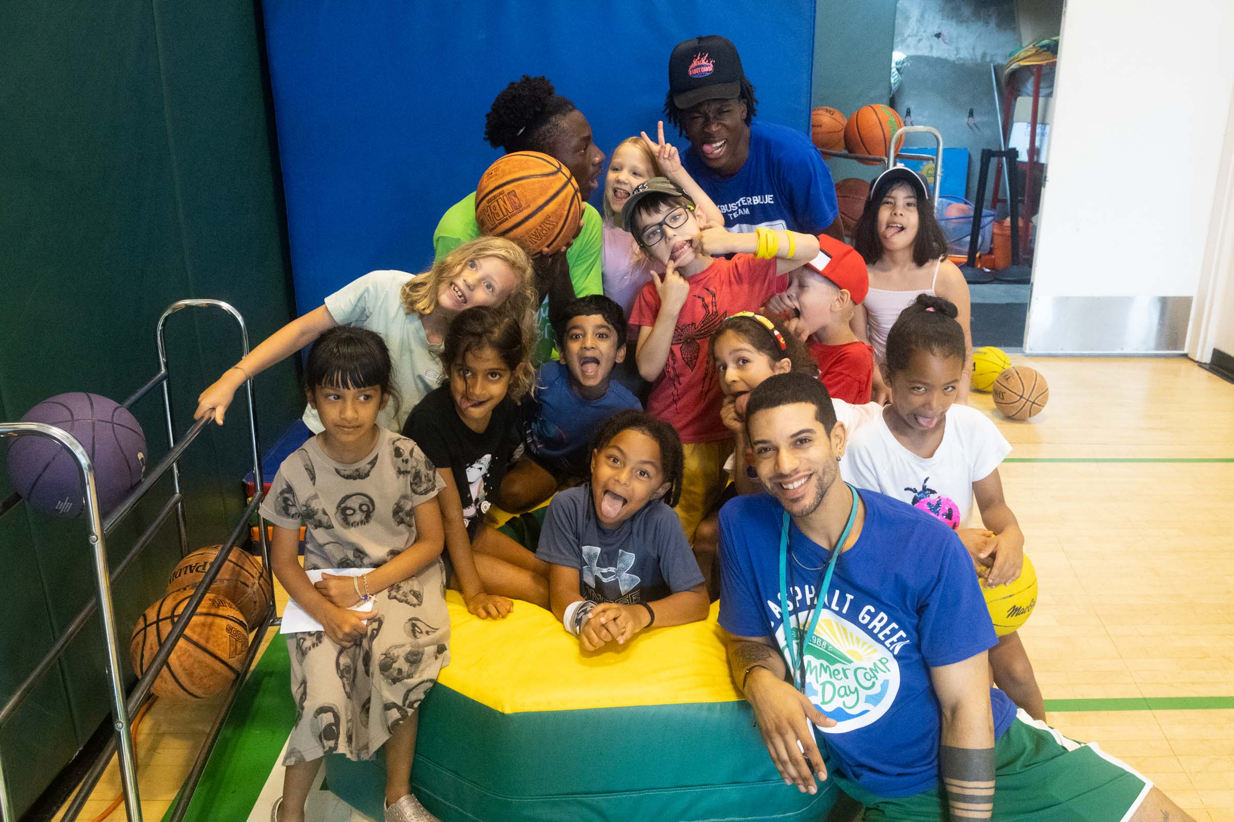 A group of kids and two adults pose for a cheerful photo in a gym, surrounded by basketballs. One adult and several children make playful expressions.