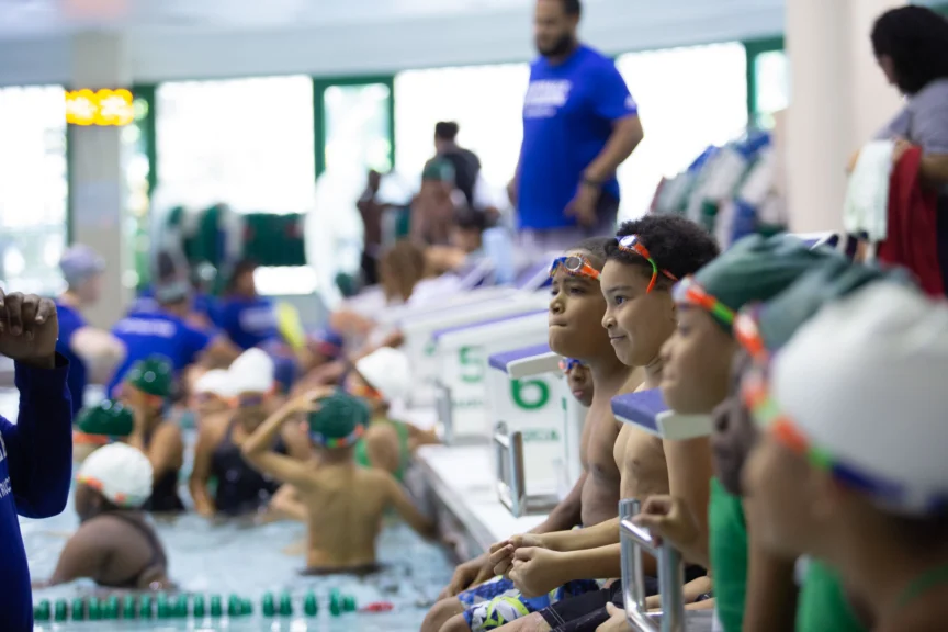 Children in swim gear sit by a pool, watching swimmers in the water. A coach in a blue shirt stands in the background.