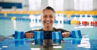 A smiling person standing in a swimming pool, holding blue foam dumbbells.