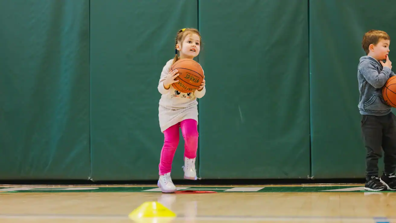 A young girl in pink leggings jumps while holding a basketball, with a boy nearby also ready for youth basketball practice in a gym with green padded walls.