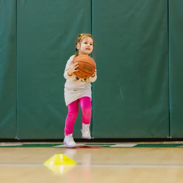 A young girl in pink leggings jumps while holding a basketball, with a boy nearby also ready for youth basketball practice in a gym with green padded walls.
