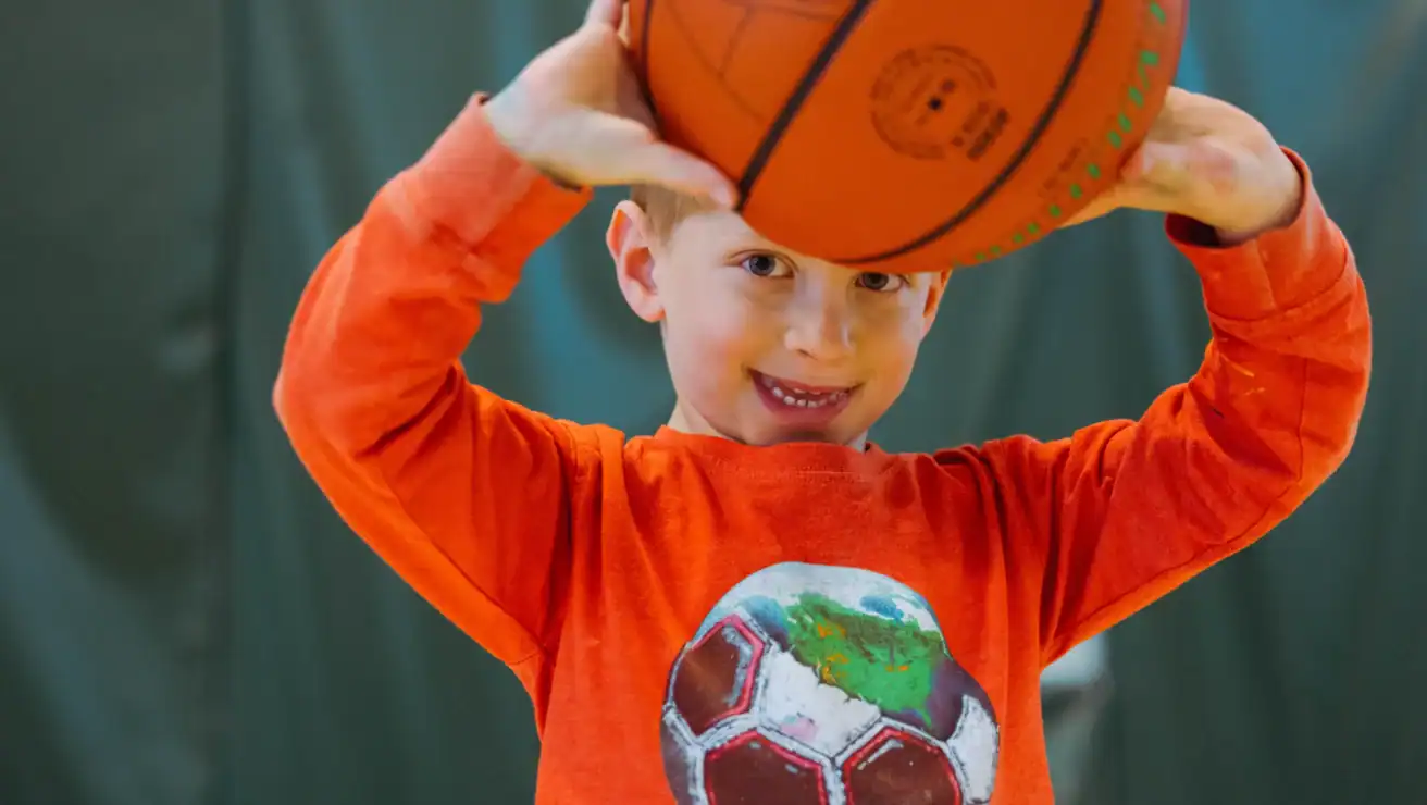 Young boy in an orange shirt with a soccer ball graphic holds a basketball above his head, smiling at the camera, ready for youth basketball fun.