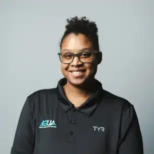 A person wearing glasses and a black collared shirt with ASUA and TYR logos stands smiling in front of a plain gray background.