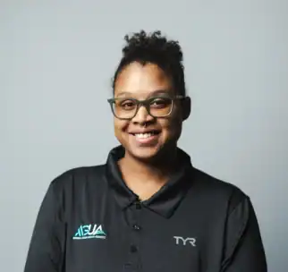 A person wearing glasses and a black collared shirt with ASUA and TYR logos stands smiling in front of a plain gray background.