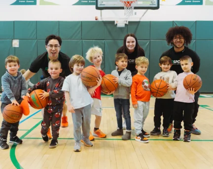 A group of young children holding basketballs poses with three adult coaches an Asphalt Green Youth Basketball Class.