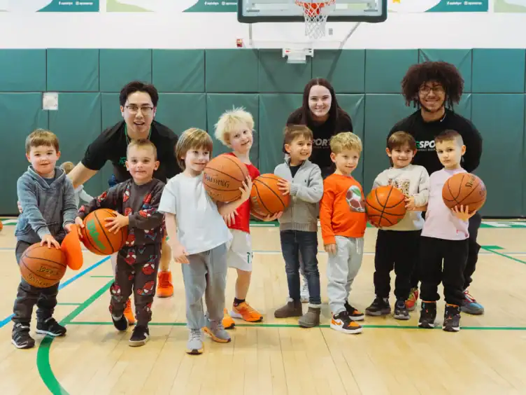 A group of young children holding basketballs poses with three adult coaches on an indoor youth basketball court.