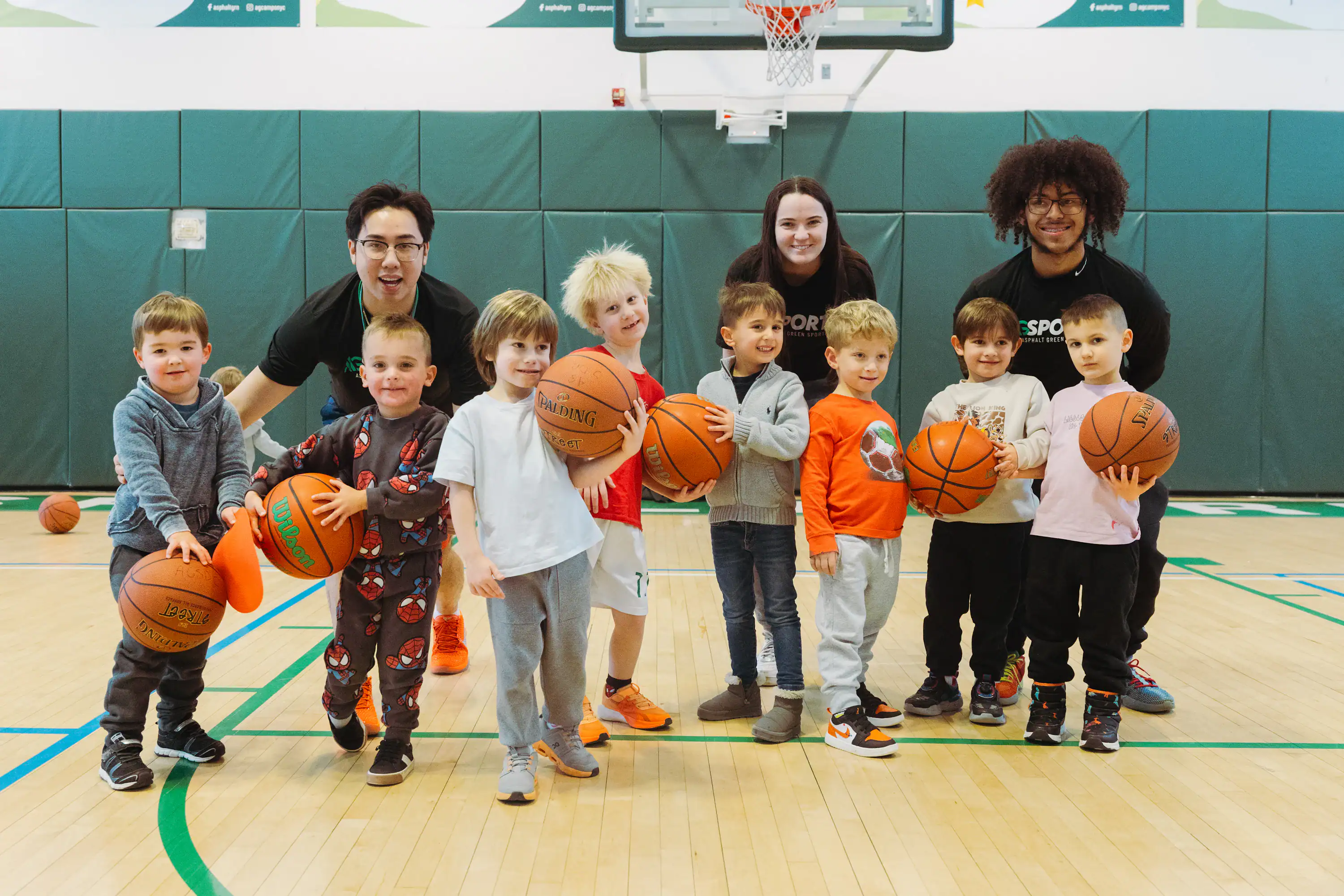 A group of young children holding basketballs poses with three adult coaches on an indoor youth basketball court.