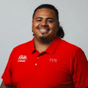 A person wearing a red lifeguard polo shirt with "ASJA Lifeguard" and "TYR" logos smiles in front of a plain light gray background.