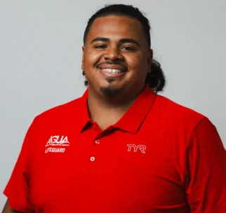 A person wearing a red lifeguard polo shirt with "ASJA Lifeguard" and "TYR" logos smiles in front of a plain light gray background.