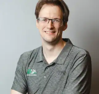 A man with glasses and light brown hair smiles at the camera, wearing a gray collared shirt with "AUSA" and "TYR" logos, standing against a plain light background.