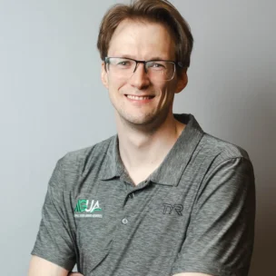 A man with glasses and light brown hair smiles at the camera, wearing a gray collared shirt with "AUSA" and "TYR" logos, standing against a plain light background.