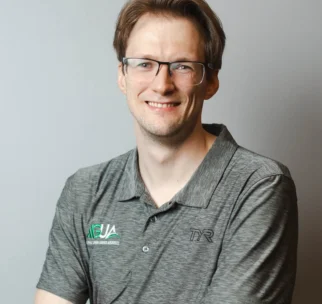 A man with glasses and light brown hair smiles at the camera, wearing a gray collared shirt with "AUSA" and "TYR" logos, standing against a plain light background.