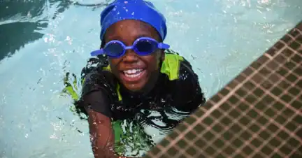 A child wearing blue googles and a blue swim cap smiling in a pool.