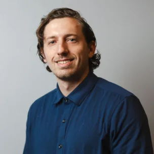 A man with wavy brown hair, wearing a dark blue button-up shirt, smiles at the camera against a plain light gray background.