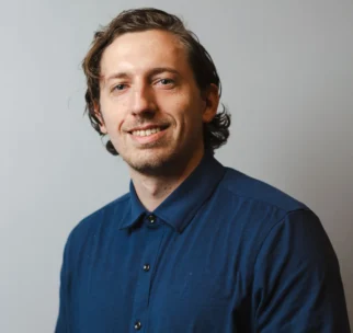 A man with wavy brown hair, wearing a dark blue button-up shirt, smiles at the camera against a plain light gray background.