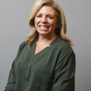A woman with shoulder-length blonde hair wearing a dark green blouse stands and smiles in front of a plain gray background.