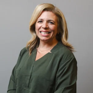 A woman with shoulder-length blonde hair wearing a dark green blouse stands and smiles in front of a plain gray background.
