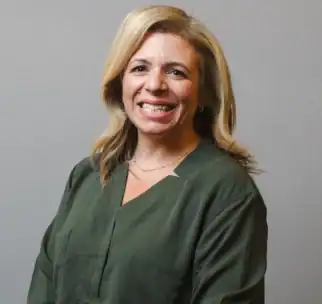 A woman with shoulder-length blonde hair wearing a dark green blouse stands and smiles in front of a plain gray background.