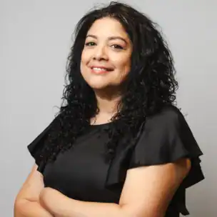 Woman with long curly black hair wearing a short-sleeved black top stands with arms crossed, smiling slightly, in front of a plain light gray background.