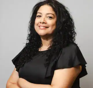 Woman with long curly black hair wearing a short-sleeved black top stands with arms crossed, smiling slightly, in front of a plain light gray background.