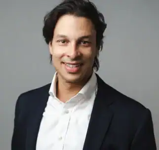 A man with dark hair wearing a white shirt and dark blazer poses for a professional portrait against a plain gray background.