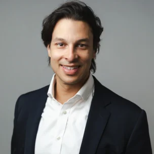 A man with dark hair wearing a white shirt and dark blazer poses for a professional portrait against a plain gray background.