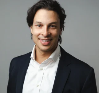 A man with dark hair wearing a white shirt and dark blazer poses for a professional portrait against a plain gray background.
