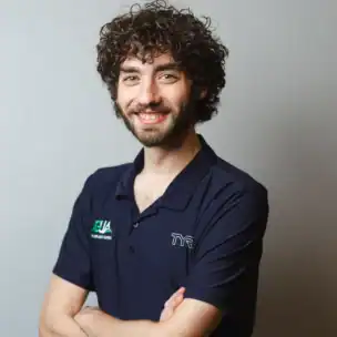 A man with curly hair and a beard, wearing a navy collared shirt with logos, stands against a plain light background with his arms crossed and smiling.