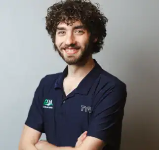 A man with curly hair and a beard, wearing a navy collared shirt with logos, stands against a plain light background with his arms crossed and smiling.