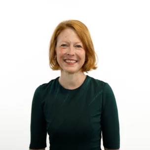 A woman with red hair wearing a dark green dress stands against a plain white background and smiles at the camera.