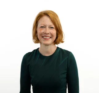 A woman with red hair wearing a dark green dress stands against a plain white background and smiles at the camera.