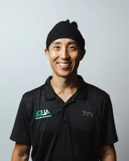 A person wearing a black collared shirt and black headband smiles at the camera against a plain light background.