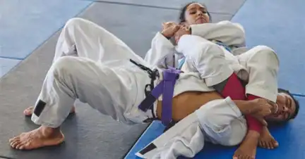 Two people in jiu-jitsu uniforms practice on mats; one performs an armbar submission while the other attempts to defend.