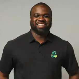 A man wearing a black polo shirt with a colorful camp logo stands smiling in front of a plain gray background.