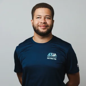 A man stands against a plain light background wearing a navy blue shirt with "Swim Academy Instructor" text on the front.