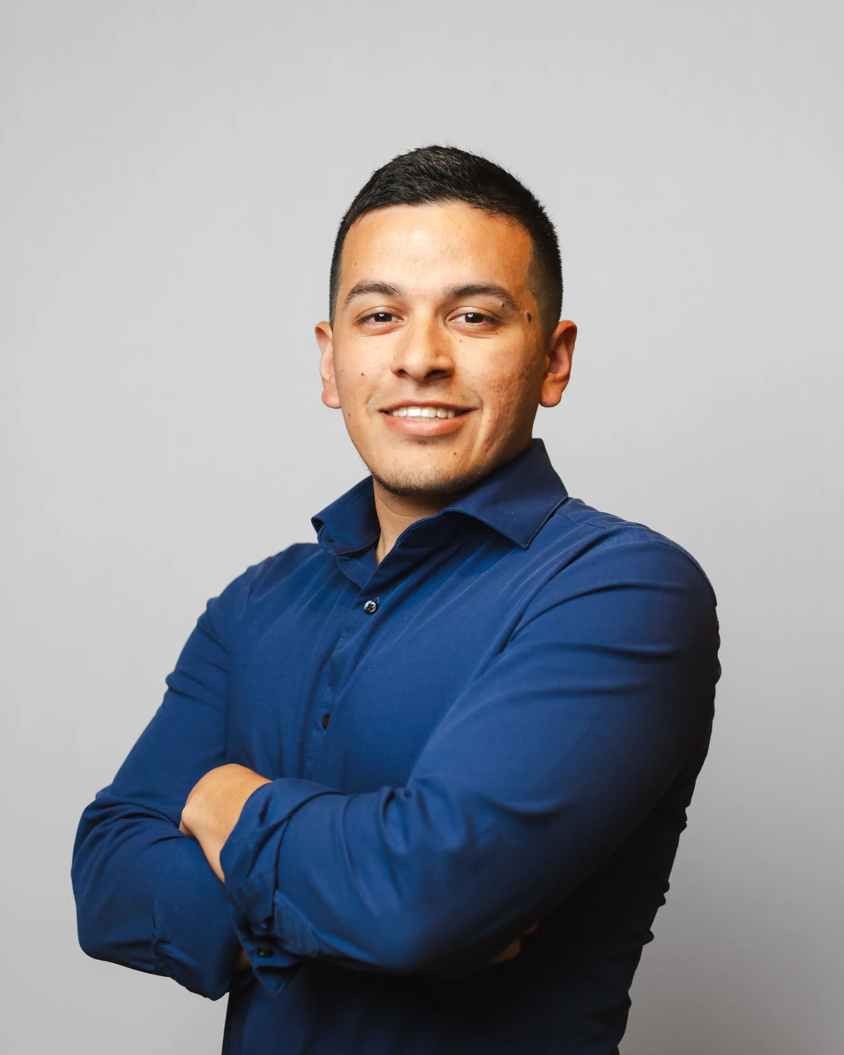 A man with short dark hair, wearing a dark blue button-up shirt, stands with arms crossed and smiles at the camera against a plain light gray background.
