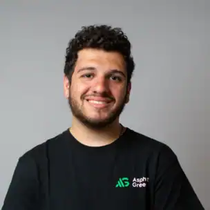 A young man with curly hair and a beard stands against a plain gray background, wearing a black Asphalt Green t-shirt and smiling at the camera.